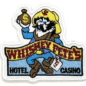 May include: An embroidered patch featuring a cartoon character holding a bag of money. The character is wearing a yellow hat and blue clothing. The patch has the text "Whiskey Pete's Hotel Casino" in red and yellow lettering.