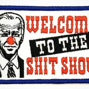 May include: Embroidered patch with a blue border, featuring a cartoon image of a man with a red nose and the text "WELCOME TO THE SHIT SHOW".