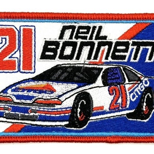 May include: A red, white, and blue embroidered patch featuring a race car with the number 21 on the side. The text "Neil Bonnett" is above the car.