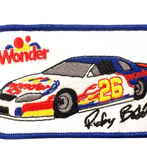 Puede incluir: Un parche rectangular bordado con fondo blanco y borde azul. Presenta un coche de carreras con colores azul, blanco, rojo y amarillo, el número 26, el logotipo de Wonder y la firma Ricky Bobby en hilo de bordar de color negro.