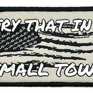 Puede incluir: Un parche bordado beige con costuras negras. El parche presenta una bandera estadounidense estilizada con el texto "TRY THAT IN A SMALL TOWN" en blanco.