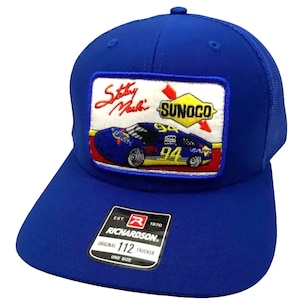 May include: A blue trucker hat with a red, yellow and white patch featuring a racing car and the text "Sunoco" and "Staley Nash's". The hat also has a black label with the text "Richardson" and "112 Trucker".