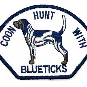 May include: White embroidered patch with a blue border. The patch features a black and white hound dog with brown markings. The text "COON HUNT WITH BLUETICKS" is embroidered in blue.