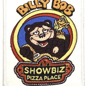 May include: Embroidered patch featuring Billy Bob, a brown bear character, from Showbiz Pizza Place. The patch has a white border and includes the text "BILLY BOB" and "SHOWBIZ PIZZA PLACE". The bear is holding a fork and is next to a yellow bird.