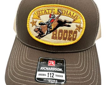 May include: Brown and beige trucker hat with a patch that says "State Finals Rodeo" with a cowboy riding a bull. The patch has a yellow border and stars on either side. The hat has a Richardson 112 label.