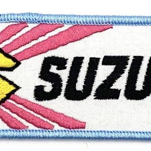 Könnte beinhalten: Ein weißer gestickter Aufnäher mit blauem Rand. Der Aufnäher zeigt ein stilisiertes gelbes "S" mit rosa Strahlen, die von ihm ausgehen. Das Wort "SUZUKI" ist in Schwarz unter dem "S" gestickt.