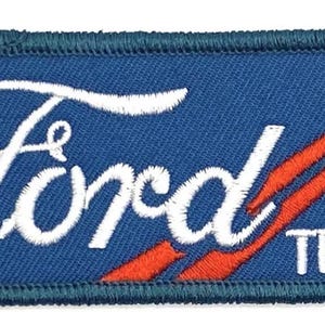 May include: A blue and white embroidered patch with the word "Ford" in a white cursive font and the word "Trucks" in white below. The patch has two red diagonal stripes in the background.