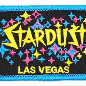 May include: Embroidered patch with the word "STARDUST" in yellow, with a variety of pink, blue, and yellow starburst designs. The text "LAS VEGAS" is below the main text. The patch has a black background and a blue stitched border.
