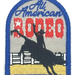 May include: An embroidered patch with a blue border, featuring a cowboy riding a bucking bull in front of a fence. The text "All American Rodeo" is above the cowboy and bull.