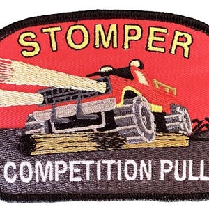May include: Embroidered patch featuring a red monster truck with large tires, emitting light beams. The word "STOMPER" is arched above the truck, and "COMPETITION PULL" is below. The patch has a black border and a red background.