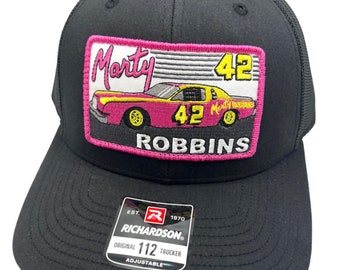 May include: Black trucker hat with a pink and yellow patch featuring a race car with the text "Marty Robbins 42" and the logo "Richardson 112 Original Trucker Adjustable".