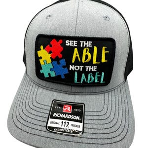 May include: A grey and black trucker hat with a black patch that says "See the Able Not the Label" with a colourful puzzle piece design. The hat has a black mesh back and an adjustable strap with a Richardson 112 label.