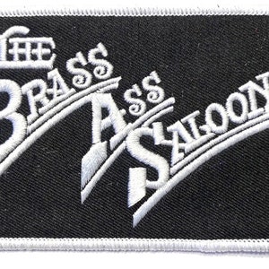 May include: White embroidered patch on a black background with the text "The Brass Saloon" in a stylized font.