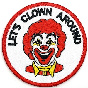 May include: A white, round embroidered patch with a red border. The patch features a cartoon clown with red hair, a white face, and a big smile. The text "Let's Clown Around" is written in a black circle around the clown.