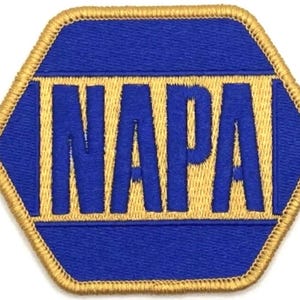 May include: A blue and gold embroidered patch in a hexagonal shape. The word "NAPA" is embroidered in gold block letters on a blue background. The patch has a gold border.