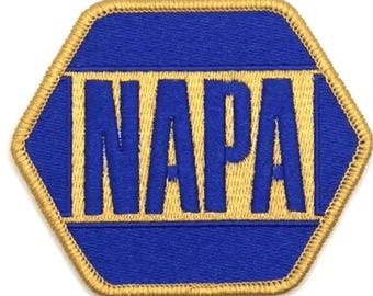 May include: A blue and gold embroidered patch in a hexagonal shape. The word "NAPA" is embroidered in gold block letters on a blue background. The patch has a gold border.