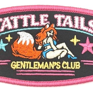 May include: Embroidered patch with a black background and a pink border. The patch features the text "TATTLE TAILS GENTLEMAN'S CLUB" and a cartoon fox woman. Stars in pink, yellow, and blue are also embroidered.