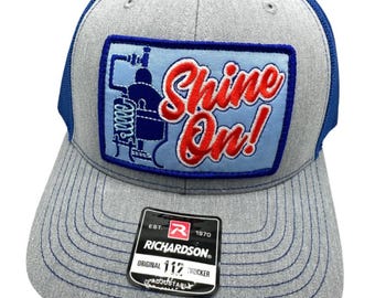May include: Gray trucker hat with a blue mesh back and brim. The front patch features a blue and red design with the words "Shine On!". The hat includes a Richardson tag.