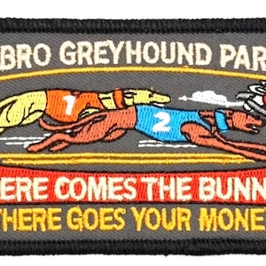 以下が含まれることがあります： 「EBRO GREYHOUND PARK」の文字と2匹のグレイハウンドが描かれた刺繍パッチ。「HERE COMES THE BUNNY - THERE GOES YOUR MONEY」のフレーズが含まれています。犬は1と2のゼッケンを着ており、機械ウサギが描かれています。