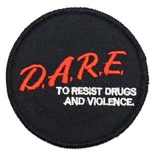 May include: A black circular patch with white stitching around the edge. The text "D.A.R.E. TO RESIST DRUGS AND VIOLENCE." is embroidered in white on the patch. The letters "D.A.R.E." are embroidered in red.
