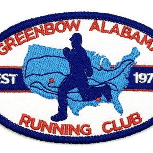 May include: A white oval patch with a blue border. The patch has a red and white banner with the text "GREENBOW ALABAMA EST 1976". A blue silhouette of a runner is superimposed on a blue and white outline of the United States. The banner below the runner reads "RUNNING CLUB".