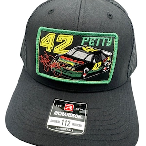 May include: Black trucker hat with a green, yellow and red embroidered patch featuring the number 42, the word "PETTY" and a race car. The patch is surrounded by a green border. The hat has a black mesh back and a label that reads "EST. 1970 RICHARDSON ORIGINAL 112 TRUCKER ADJUSTABLE".