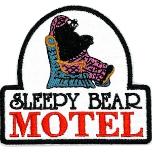 May include: Embroidered patch featuring a black bear in a pink and blue outfit, seated in a blue armchair. The patch has the words "SLEEPY BEAR MOTEL" in black and red lettering, with a black border.