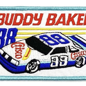 May include: A white and blue embroidered patch featuring a race car with the number 88 on it. The car is sponsored by Cisco and the patch says "Buddy Baker" at the top.
