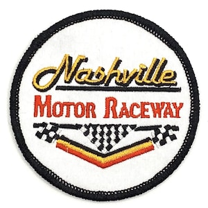 May include: A white embroidered patch with black stitching around the edges. The patch features the text "Nashville Motor Raceway" in red and yellow lettering with a checkered flag design below.
