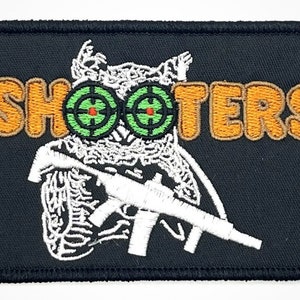 Könnte beinhalten: Ein gestickter Aufnäher mit einer weißen Eule, die ein weißes Gewehr hält. Die Augen der Eule sind grün mit roten Zielscheiben. Das Wort "SHOOTERS" ist in Orange über der Eule gestickt.