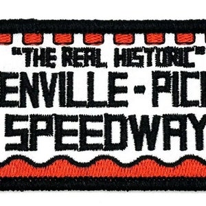 Puede incluir: Un parche blanco bordado con texto negro que dice "The Real Historic Greenville-Pickens Speedway". El parche tiene un borde ondulado rojo y dos banderas a cuadros blancas y negras a cada lado.