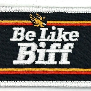 May include: Embroidered patch with the text "Be Like Biff" in white, stylized lettering on a black background. The patch features a white border and a design of a yellow eagle above the text. Red and yellow stripes are at the top and bottom.