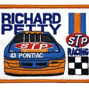 Könnte beinhalten: Ein weißer gestickter Aufnäher mit dem Text "RICHARD PETTY" in blauer Schrift. Der Aufnäher zeigt ein blaues und orangefarbenes Rennfahrzeug mit der Nummer "43" und dem Text "PONTIAC" auf der Vorderseite. Das Fahrzeug hat einen roten Kreis mit dem Text "STP" auf der Motorhaube. Der Aufnäher enthält auch einen roten Kreis mit dem Text "STP" und dem Text "RACING" darunter. Eine karierte Flagge befindet sich in der unteren rechten Ecke.