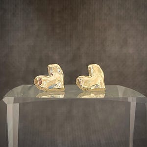 May include: A pair of gold heart-shaped earrings. The earrings are made of a textured metal and have a slightly irregular shape.