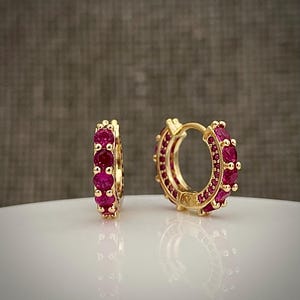 May include: A pair of gold hoop earrings featuring a row of round, vibrant pink stones set along the front edge. The earrings have a hinged closure mechanism and a polished metallic finish, reflecting light on the smooth surface below.