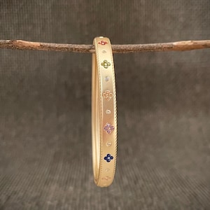 Op de afbeelding: Een goudkleurige armband met een bloemmotief. De armband heeft kleine, kleurrijke edelstenen in rood, groen, roze en blauw en wordt gepresenteerd op een houten tak tegen een neutrale achtergrond.