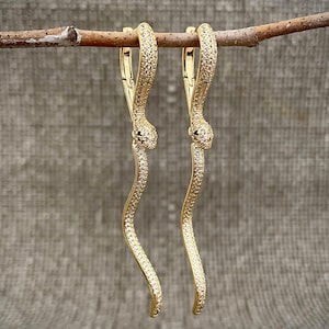 May include: A pair of gold-toned drop earrings shaped like elongated snakes. Each earring features a hinged hoop top and a long, curved body encrusted with small, clear pavé stones. The snake heads are detailed with tiny black eyes, creating a symmetrical design.