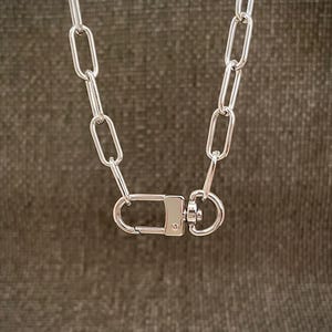 May include: A silver-colored chain necklace with large, rectangular links. The necklace features a clasp closure with a rectangular bar and a circular ring. The background is a solid, neutral color.
