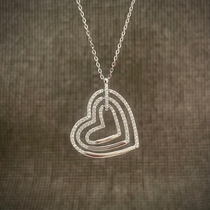 Può includere: Una collana d'argento con un ciondolo a forma di tre cuori intrecciati, ognuno adornato con scintillanti pietre di zircone cubico trasparenti.
