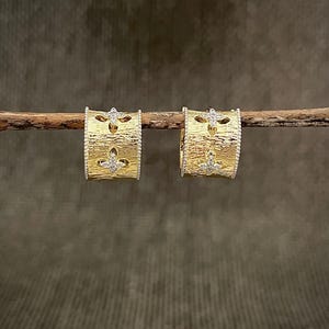 Könnte beinhalten: Ein Paar goldfarbene Creolen mit strukturierter Oberfläche. Jeder Ohrring hat ein Fleur-de-Lis-Design mit kleinen, klaren Edelsteinen. Die Ohrringe werden auf einem Holzast vor neutralem Hintergrund präsentiert.