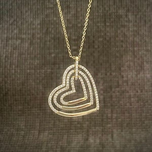 Può includere: Una collana d'oro con un ciondolo a forma di tre cuori intrecciati, ognuno adornato con pietre chiare scintillanti.