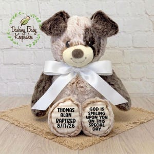 May include: A plush puppy dog with brown and white fur, a white bow, and embroidered foot pads. The foot pads read "THOMAS ALAN BAPTIZED 8/17/26" and "GOD IS SMILING UPON YOU ON THIS SPECIAL DAY".