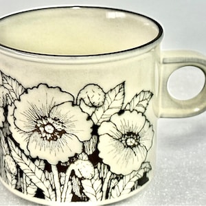 May include: A white ceramic mug with a black and brown floral design. The mug has a rounded handle and a black rim.