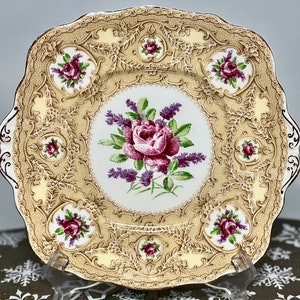 Op de afbeelding: Een vierkant, goudkleurig, keramisch bord met een delicaat bloemenontwerp. Het bord heeft een centraal boeket van roze rozen en paarse lavendel, omringd door een herhalend patroon van kleinere rozen en ingewikkelde krulwerk.