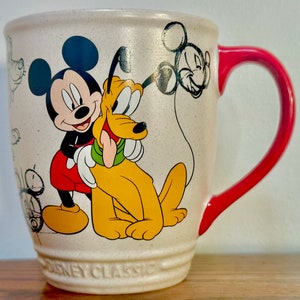 May include: A white ceramic mug with a red handle features a cartoon illustration of Mickey Mouse and Pluto. Mickey is wearing red shorts and Pluto is yellow. The mug has the text "Disney Classic" printed on the bottom.