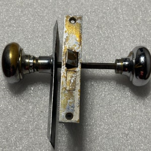 May include: A vintage chrome door knob and latch set with a brass accent. The knob is round and has a smooth surface. The latch is rectangular and has a keyhole.