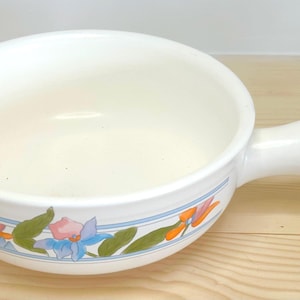 May include: White ceramic bowl with a handle, featuring a floral design in orange, blue, and green. The bowl has a rounded shape and a single handle. The floral pattern is on a blue stripe around the rim. A kitchenware item.