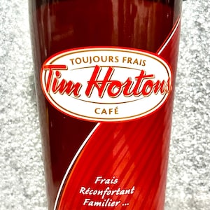 Peut inclure: Un gobelet à café Tim Hortons rouge et brun avec un couvercle brun. Le gobelet porte le logo Tim Hortons et le texte "Toujours Frais" et "Café". Le gobelet porte également le texte "Frais Reconfortant Familier... Toujours" et "Depuis 1964".