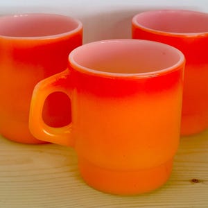Puede incluir: Tres tazas de vidrio naranja con un borde blanco. Las tazas son degradadas, con un naranja más claro en la parte superior y un naranja más oscuro en la parte inferior.