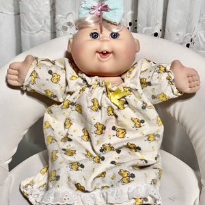 Puede incluir: Una muñeca Cabbage Patch Kid con un camisón de estampado floral blanco y amarillo con una cinta amarilla y un lazo azul claro en la cabeza.